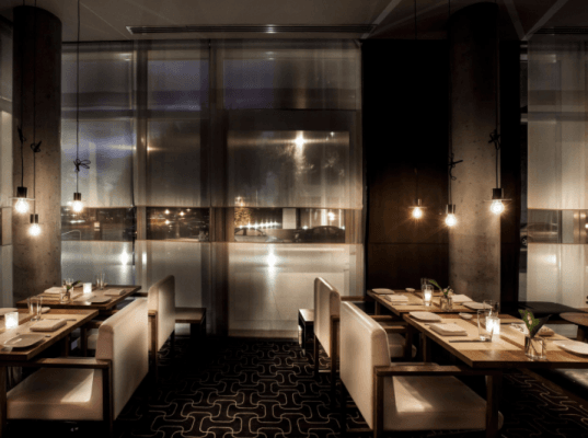 A modern, dimly lit restaurant interior with white cushioned chairs, wooden tables set for Pizza Suprema dining, pendant lights hanging above, and large frosted windows creating a cozy atmosphere.
